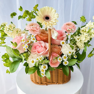 A floral arrangement featuring a variety of flowers in pink, yellow, and white, nestled in a wicker basket against a white background.