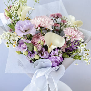 A floral bouquet featuring a variety of flowers including purple and white lilies, chrysanthemums, and other mixed flowers, tied with a ribbon and presented in a clear wrapper.