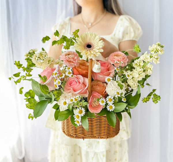 A floral arrangement featuring a variety of flowers in pink, yellow, and white, nestled in a wicker basket, send a basket of joy