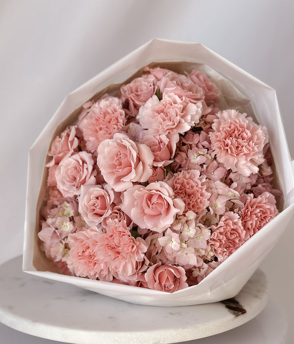 A round bouquet of pink hydrangeas, spray roses, and carnations arranged in a white container, wrapped in luxury paper and tied with a satin ribbon.