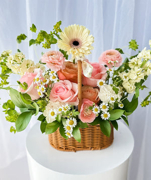 A floral arrangement featuring a variety of flowers in pink, yellow, and white, nestled in a wicker basket against a white background.