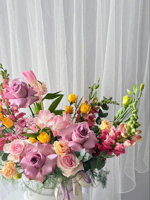 Colorful flower arrangement in a white box with a purple ribbon against a white curtain background