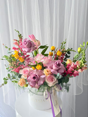 Colorful flower arrangement in a white box with a visible brand name against a white curtain background.