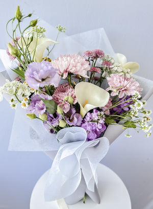 A floral bouquet featuring a variety of flowers including purple and white lilies, chrysanthemums, and other mixed flowers, tied with a ribbon and presented in a clear wrapper.