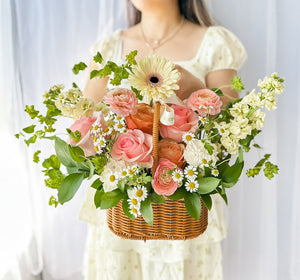 A floral arrangement featuring a variety of flowers in pink, yellow, and white, nestled in a wicker basket, send a basket of joy