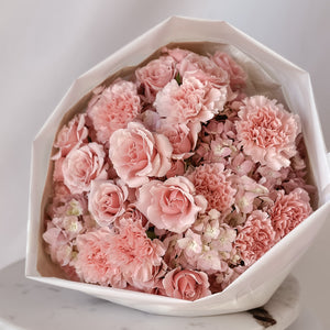 A round bouquet of pink hydrangeas, spray roses, and carnations arranged in a white container, wrapped in luxury paper and tied with a satin ribbon.