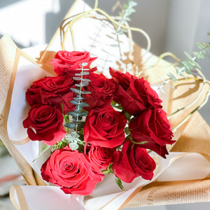 A bouquet of red roses with green eucalyptus leaves and curly willow, wrapped in white paper with a ribbon.