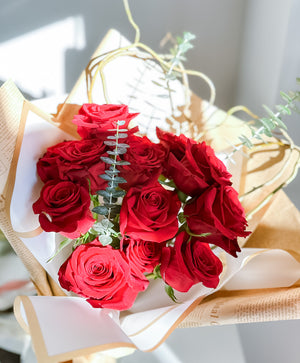 A bouquet of red roses with green eucalyptus leaves and curly willow, wrapped in white paper with a ribbon.